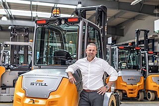 Muž v bílé košili se opírá o oranžový vysokozdvižný vozík s logem „STILL“ v hale, v pozadí jsou další vysokozdvižné vozíky